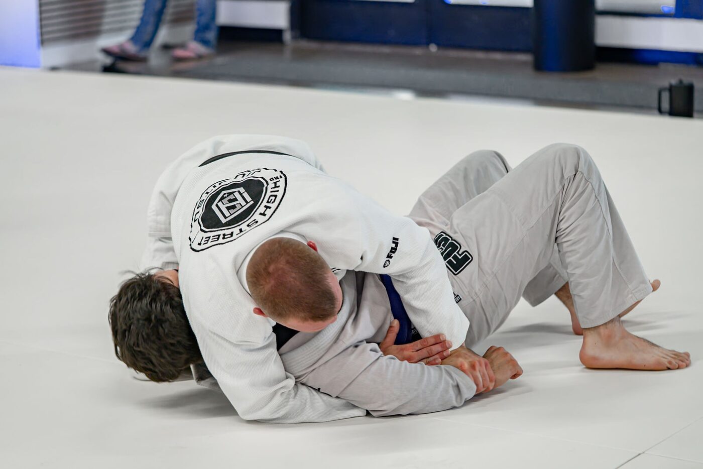 Two adults in white gis grapple on the mats, one working from turtle while the other controls the back